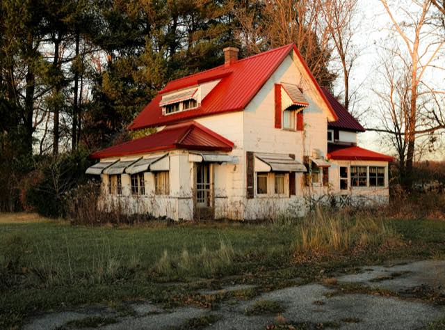 Older white house with a red metal roof, worn siding and an overgrown yard